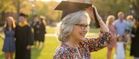 Happy mature Caucasian woman in graduation cap celebrating academic achievement outdoors with warm sunlight and blurred crowd background for education success.