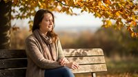 Young woman relaxing on wooden bench under autumn tree with golden leaves, enjoying peaceful moment outdoors in warm sunlight during fall season.