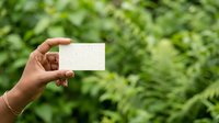 Hand holding blank white card against lush green foliage background, suitable for business card mockups, eco-friendly branding, and nature-themed marketing materials.
