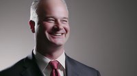 Confident mature Caucasian businessman in dark suit and red tie smiling warmly against studio background, professional corporate portrait with dramatic lighting.