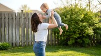 Young mother lifting happy baby boy in air during outdoor playtime in sunny backyard garden with wooden fence and green grass creating joyful family bonding moment.