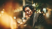 Middle-aged Asian man with curly hair smiling warmly at camera in cozy evening setting with golden bokeh lights creating intimate atmospheric mood for lifestyle content.