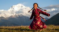 Young Asian woman in traditional red dress dancing joyfully in mountain meadow with snow-capped peaks and dramatic sky at sunset.