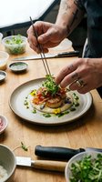Chef with tattooed arms plating gourmet dish with microgreens and chopsticks on wooden counter, fine dining preparation and food styling.