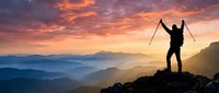 Triumphant hiker silhouette celebrating on mountain peak at sunrise with trekking poles raised against dramatic orange purple sky and layered mountain ranges.