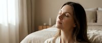 Young woman with closed eyes relaxing peacefully on bed in bright bedroom, enjoying morning sunlight and tranquil moment of meditation and wellness.