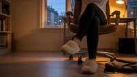 Woman in black leggings and fuzzy slippers relaxing on office chair at home desk during evening, feet up, cozy workspace lighting.
