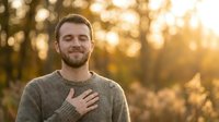 Young Caucasian man with beard wearing gray sweater, hand on chest, smiling with eyes closed in peaceful autumn forest setting during golden hour.