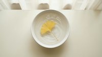 Single yellow ginkgo leaf floating in white ceramic bowl with water on light wooden surface for zen meditation and natural spa wellness concepts.
