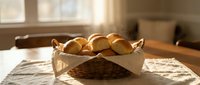 Fresh baked dinner rolls in wicker basket with white cloth on wooden table in warm sunlit kitchen for home cooking and baking concepts.
