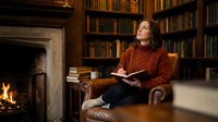 Young woman reading book in cozy library with fireplace, relaxing in leather armchair surrounded by bookshelves, warm autumn evening atmosphere.