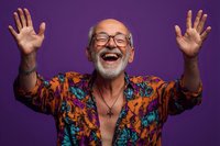 Joyful senior man with gray beard and glasses celebrating with raised hands wearing colorful tropical shirt on purple background.