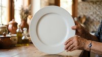 Senior woman with brown skin holding empty white plate in rustic kitchen with pottery and natural light, representing food service, cooking, meal preparation, and culinary themes.
