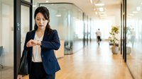 Young Asian woman in navy blazer checking wristwatch in modern office corridor with glass partitions, representing time management and professional business environment.