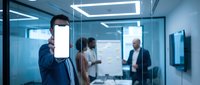 Professional businessman in suit holding blank smartphone screen in modern office conference room with colleagues in background meeting.