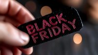 Pink neon glowing Black Friday text on black paper tag held by fingers against blurred bokeh lights background for seasonal sale promotions.