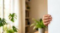 Close-up of male hand peeking around white wall corner in bright interior space with potted green plants, suggesting curiosity, surprise or playful peek. For advertising and editorial use.