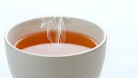 Hot steaming tea in white ceramic cup with vapor rising from amber liquid surface on clean background for beverage and relaxation concepts.