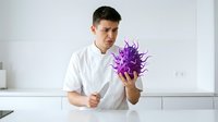 Young man with dark hair and olive complexion wearing white shirt holding purple spiky virus model in modern kitchen setting for educational or scientific presentation purposes.