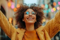 Young African American woman with natural curly hair and glasses smiling joyfully, wearing yellow teddy coat and orange sweater against blurred festive bokeh lights.