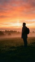 Silhouetted male hiker with backpack stands in misty meadow at sunrise, gazing at golden sky and distant forest. Ideal for travel, adventure, and outdoor lifestyle concepts.