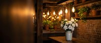 Cozy rustic cafe interior with exposed brick wall, wooden shelves, hanging Edison bulb lights, white flowers in metal bucket, and warm ambient lighting.