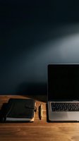 Moody dark workspace with open laptop, leather notebook and gold pen on wooden desk, dark background with dramatic lighting.