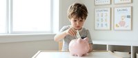 Young boy with light brown hair, Caucasian ethnicity, inserting coin into pink piggy bank at table by window in bright classroom setting. Financial literacy education.