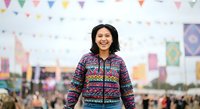 Young woman with dark hair and South Asian ethnicity wearing colorful patterned zip-up jacket at outdoor festival with bunting decorations and crowd in background.