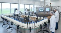 Two technicians in white lab coats monitoring automated bottling line with milk bottles in modern dairy processing facility with large windows.
