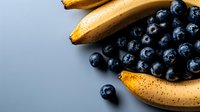 Fresh ripe blueberries scattered near spotted yellow bananas on blue background, healthy breakfast ingredients for smoothie or dessert preparation.