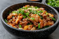 Spicy stir-fried meat and potato chunks garnished with fresh green onions and sesame seeds in black ceramic bowl, served on gray stone surface.