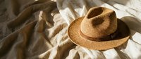 Woven straw fedora hat with brown leather band resting on crumpled white linen bedding in warm natural sunlight creating soft shadows for summer fashion.