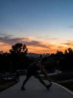 Woman in athletic wear performing a forward fold stretch on an urban park pathway at sunset, with city skyline and trees silhouetted against golden sky, ideal for fitness and wellness content.