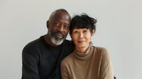 Senior African American man with gray beard and Asian woman with short dark hair embracing against neutral background, portraying love and companionship in mature interracial relationship.