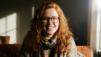 Happy young Caucasian woman with curly red hair and glasses smiling warmly in cozy indoor setting with natural sunlight streaming through window.