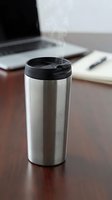 Stainless steel travel mug with steam on wooden desk next to laptop and notebook for office coffee break and workplace productivity.