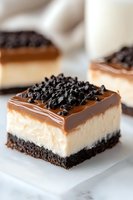 Layered cheesecake bar with chocolate cookie base, creamy vanilla filling, caramel topping and crushed cookie crumbs garnish on white marble surface. Close up food photography.