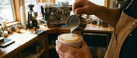 Barista pouring steamed milk into coffee cup creating latte art in cozy cafe kitchen with professional espresso machine and warm lighting atmosphere.