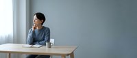 Young Asian businesswoman in casual blue sweater sitting at wooden desk in modern office, looking thoughtfully out window during work break.