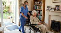 Female healthcare worker in blue scrubs assisting elderly Caucasian woman in wheelchair at comfortable home setting with bookshelf and fireplace.