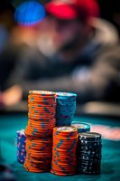 Stacks of colorful poker chips on green felt table with blurred player in background, dramatic lighting highlights orange and blue gaming pieces in casino setting.