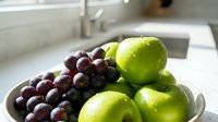 Fresh green apples and dark grapes arranged in a white bowl on a kitchen counter, natural light highlighting vibrant colors and textures.