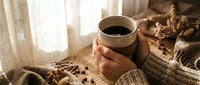 Cozy morning scene with hands holding warm coffee mug near window with soft natural light, knitted sweater and autumn decor creating hygge atmosphere.