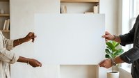 Diverse male and female professionals holding blank white poster board for custom messaging or branding in modern office setting with shelving and plant.