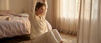 Caucasian woman in her thirties sitting on bedroom floor wearing cream pajamas, morning sunlight streaming through sheer curtains, contemplative pose for wellness and lifestyle content.