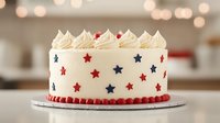 Patriotic white frosted cake decorated with red and blue stars and piped border details, topped with swirled buttercream peaks on blurred festive background.