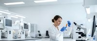 Female scientist in white lab coat examining test tube in modern laboratory with microscope and equipment for research analysis.