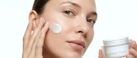 Young woman applying white facial cream moisturizer to cheek while holding cosmetic jar. Natural skincare routine and beauty treatment concept.