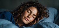 Young mixed-race woman with curly hair sleeping peacefully in bed with blue bedding, eyes closed in comfortable rest for wellness and sleep concepts.
